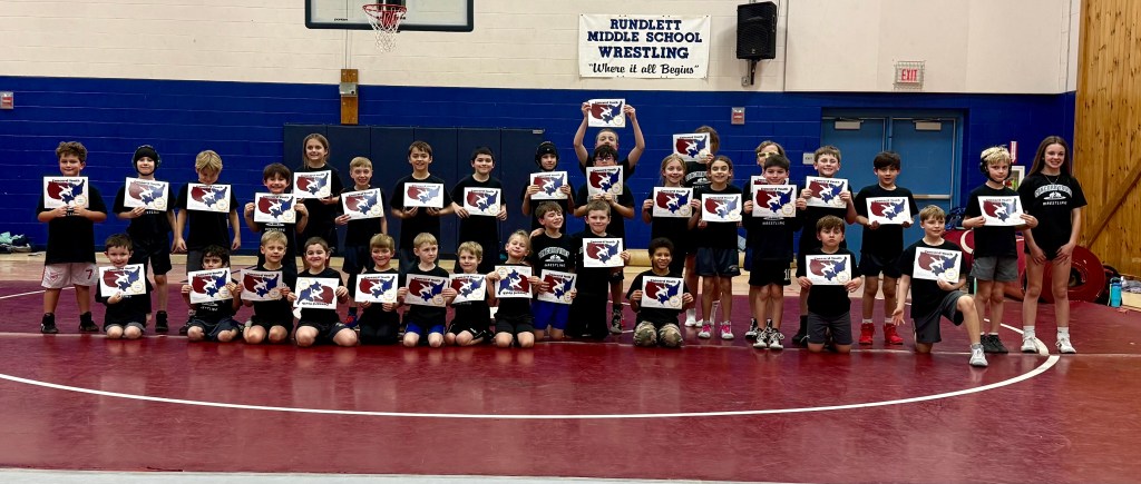 A group of children proudly holding up their wrestling certificates, standing on a red mat in a gymnasium with blue walls ...